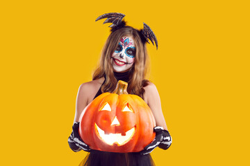 Child celebrating Halloween. Happy smiling girl in spooky skeleton costume, with Catrina skull makeup and cute bat wings on her head holding carved Jack-o-lantern pumpkin isolated on yellow background