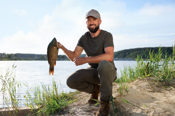 Smiling fisherman holding caught fish near lake at summer