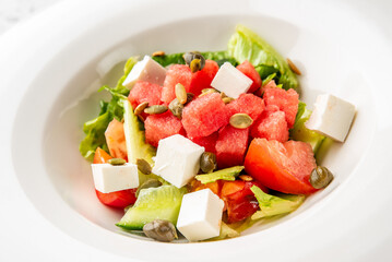 Fresh summer salad with watermelon, tomato, cucumber, lettuce, feta cheese, capers, and pumpkin seeds served in a white bowl. Colorful, healthy, and appetizing.