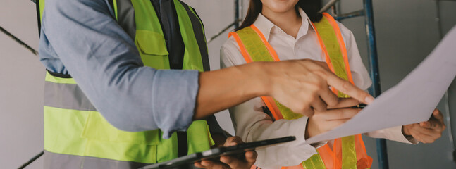 Construction manager and engineer dressed in orange work vests and hard helmets explore construction documentation on the building site near the steel frames