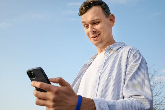 Young man smiling and looking at smartphone outdoors on a sunny day. Concept of communication, social media, positive emotions and modern lifestyle.