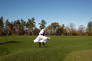 Romantic moment of a man carrying a woman in a white dress across a sunny green field with trees in...