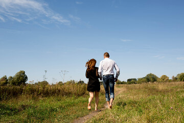Back view of a couple walking barefoot along a grassy path in a summer meadow under a blue sky.