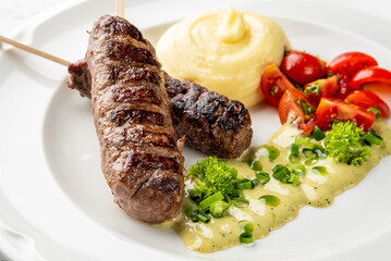 Grilled meat skewers with mashed potatoes, cherry tomatoes, and creamy herb sauce garnished with green onions and parsley on a white plate