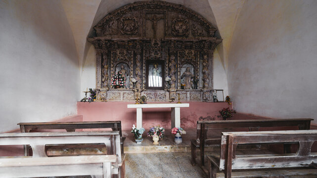 Small rustic chapel interior with wooden benches, simple altar, colorful flowers, and ornate carved religious altarpiece with saints and crucifix under arched ceiling