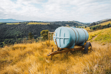 Old blue water tank trailer standing on dry yellow grass in countryside field with scenic hills and...