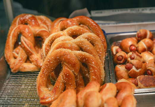 Golden-brown pretzels are showcased on a wire rack, their surfaces glistening with salt. Next to them, a tray filled with freshly baked pigs in blankets adds to the inviting bakery display. 