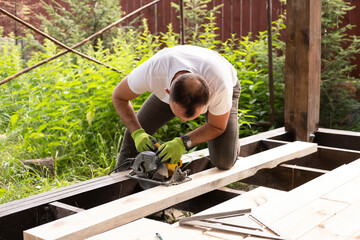 Carpenter sawing wood with an electric circular saw on sunny day at construction site.