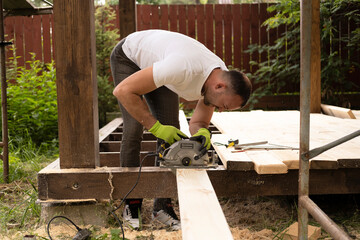 Carpenter sawing wood with an electric circular saw on sunny day at construction site.