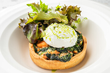 Vegetable tart with spinach, sweet potato, and poached egg, garnished with leafy greens and green seasoning, served on a white plate.