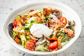 Fresh salad with mixed greens, cherry tomatoes, crispy bacon, fried onions, sesame seeds, poached egg, and creamy dressing in a white bowl on a light stone surface.