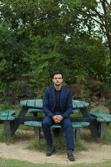 Fototapeta premium Sophisticated Man in Nature's Embrace: A well-dressed man sits serenely on a wooden bench amidst a vibrant green landscape.