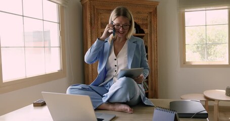 Funny professional woman in suit working in office seated cross-legged on workplace desk with laptop and tablet while lead business conversation with client on phone. Workflow, deadline, multitasking - Powered by Adobe