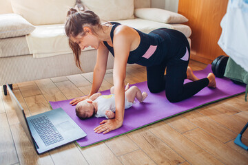 Postpartum recovery exercise at home. Young mother training together with baby girl on yoga mat using online course on laptop