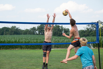 Shirtless Volleyball player hitting the ball against a block during a doubles volleyball game by a corn field 