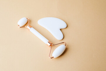 White jade roller and gua sha massage tool placed on beige surface under warm daylight for skincare...