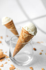 Waffle cone with vanilla ice cream ball placed in clear glass on white surface with scattered crumbs in sunlight