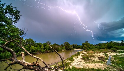 Dramatic lightning storm illuminates a riverbank fringed with lush greenery and deadwood branches.