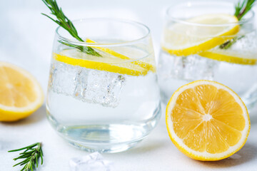 Clear glass of lemon water with rosemary sprig and ice cube beside fresh lemon half on light background in sunlight