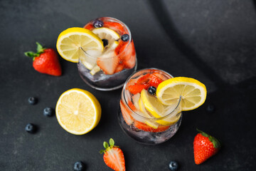 Lemonade glasses with lemon strawberries and blueberries on dark surface with scattered fruit pieces