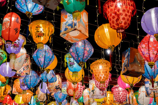 A vibrant array of lanterns, each with a unique design and color, hang outside. They are illuminated by warm yellow string lights, creating a festive atmosphere.