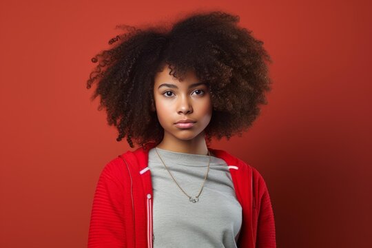 Portrait of a serious young woman with afro hair wearing red jacket and gray t shirt, posing on a red background - Powered by Adobe