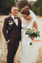 Bride and groom standing by the water, smiling and embracing on their wedding day. Elegant bridal...