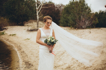 Beautiful bride with a bouquet posing by the water, her veil flowing in the wind, creating a tender...
