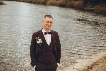 Groom standing by the water, dressed in an elegant dark suit with a bow tie and boutonniere, smiling confidently on his wedding day.