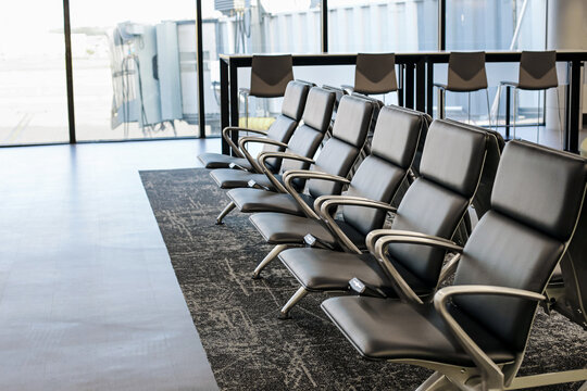 Empty black leather chairs in modern airport terminal lounge. Row of waiting seats with armrests near large glass windows. Travel concept with empty passenger area. Interior of international departure