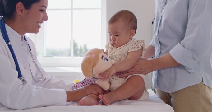Smiling pediatrician distracts an infant with a toy puppet while a parent supports during a routine checkup in a sunlit clinic. Doctor engages child with play therapy. Calm care eases fear.