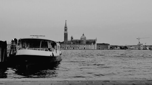 Black and white waterfront scene featuring a docked boat and a historic building with a bell tower and dome. A calm, rippled water surface adds serenity to this timeless European cityscape - Powered by Adobe