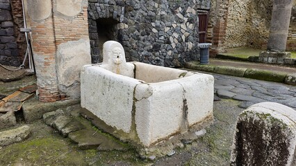 Herculaneum, Italy - 9 January 2025. A carved stone street fountain with a human face spout,...