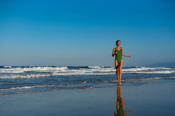 Smiling young woman in a green swimsuit walking through shallow ocean waves on a sunny beach,...