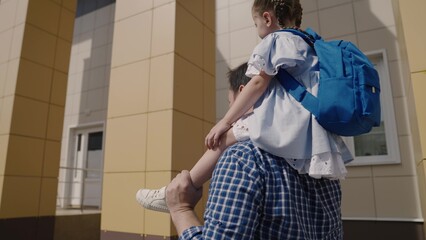 Dad carries on his shoulders little kid with backpack to school, girl with school bag rides her...