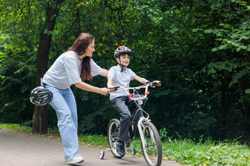 Mother teaching young son to ride bicycle with training wheels in park, family support, maternal...