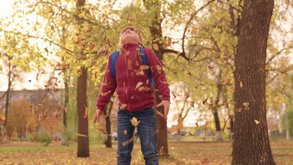 Obraz premium little happy girl with a backpack throws leaves high up and laughs cheerfully, child play in the autumn park, kid walks in the city park, raise her face looking at the golden leaf fall, autumn season.