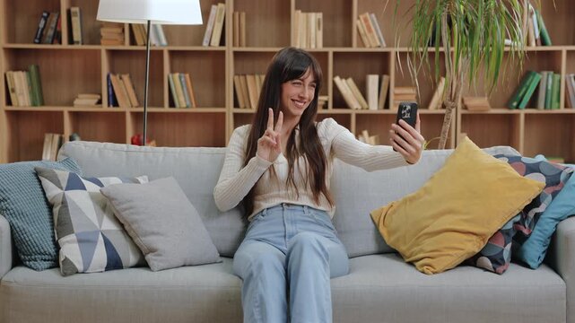Positive Caucasian woman sitting on sofa smiling at smartphone and raising fingers in peace gesture. Capturing cheerful moment or streaming video in bright living room with books and soft pillows.