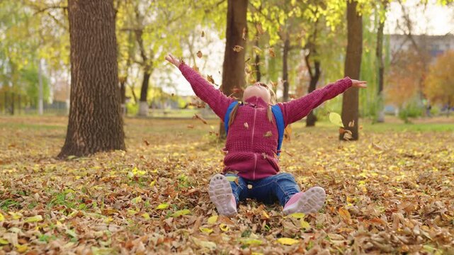 happy girl throws autumn yellow leaves up and laughs, kid is having fun playing in city nature park outdoors, little child with school backpack sits under leaf fall, first grader school yard having. - Powered by Adobe