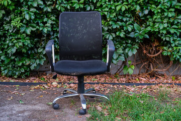 Worn black office chair with mesh backrest and armrests, left outdoors against green foliage.