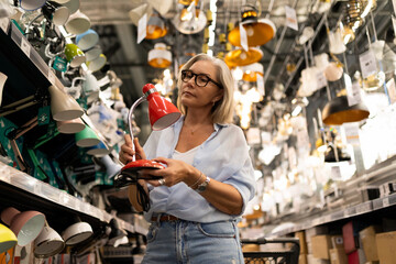 Woman shopping for a red desk lamp in a modern lighting store filled with various fixtures and...