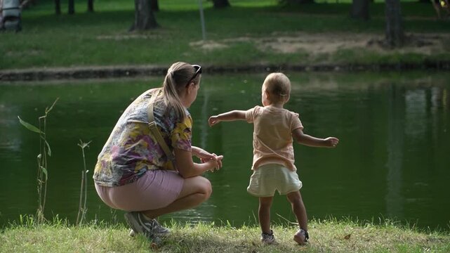girl watches attentively, sitting next to a child, as she points her finger at birds and fish in the water, enjoying the scenery and natural beauty green lake in a central city park in the summer