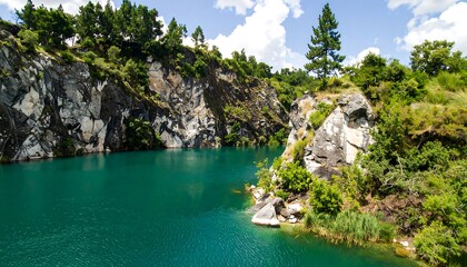 A tranquil quarry lake surrounded by dramatic, textured rock faces, vibrant greenery, and a clear, teal-colored water body under a partly cloudy sky.