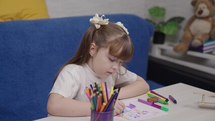 Young girl is sitting at a table with a drawing pad and a cup of markers. She is focused on her drawing, which appears to be a colorful and imaginative scene. Concept of creativity and playfulness