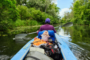 Child lies playfully in kayak while adult paddles on calm river surrounded by lush green forest....