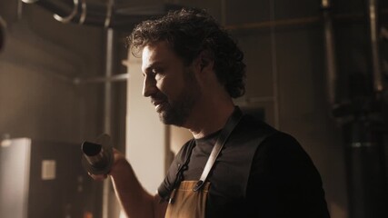 Smiling handsome Caucasian man with curly hair and beard holding metal coffee roaster close to his face. Enjoying aroma of freshly ground beans in warm coffee production room. Proud of product.