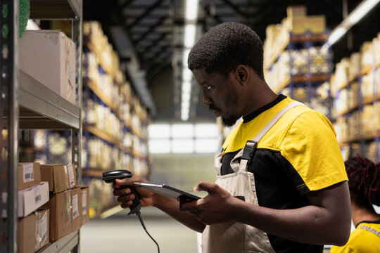 Black staff member using a barcode scanner to process shipping labels on boxes from industrial storage racks. Scanning awb tags for package tracking service with tracking info, accurate logistics.