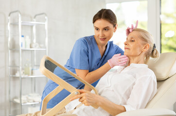 Fototapeta premium Young female doctor consulting elderly woman patient lying on clinical chair in beauty clinic