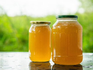 Two glass jars of golden bee honey are on a wet table, green grass field and sky in the background. Fine organic product concept. Agriculture and food industry.