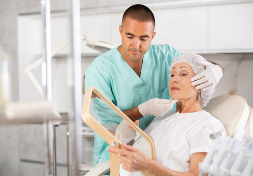 Male plastic surgeon giving professional advice after facial contouring to senior woman client looking at mirror in clinic - Powered by Adobe